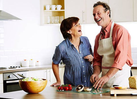 Photo of a couple cooking together in a kitchen with bowl and food on the counter. They are looking at each other laughing and smiling.