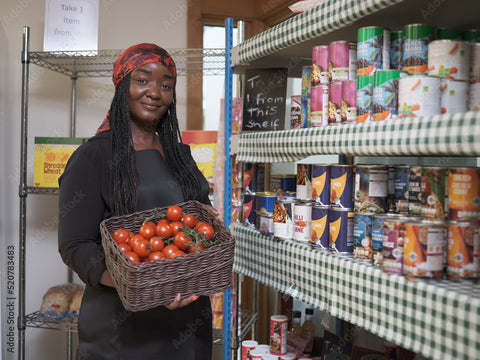 Image of a black lady holding a box of food, standing next to a block of shelves full of food baskets.
