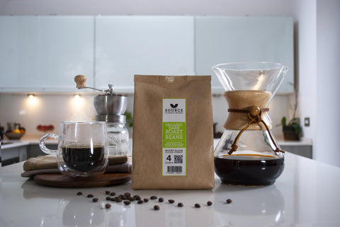 Image of Source Coffee packet. The bag is sitting on the table with a coffee pot to the right of it and a glass coffee cup to the left of it. Coffee beans are scattered across the table.