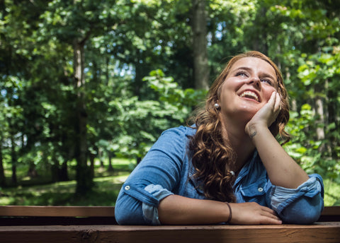 A young woman sitting on a park bench, staring up at the sky smiling. She is wearing a blue jean jacket and there is a backdrop of lush and vibrant greenery behind her.