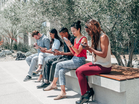 Image of multiple people, sitting in a line, on their smartphones. They are sitting outside, on a concrete bench, with trees in the background.