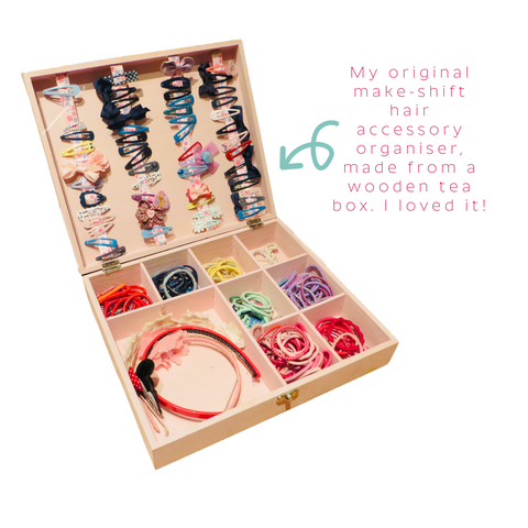 The original hair accessory box made from a wooden tea box, painted in pink, with ribbons attached inside the lid and compartments for hair bobbles. It is filled with accessories and is shown against a white background