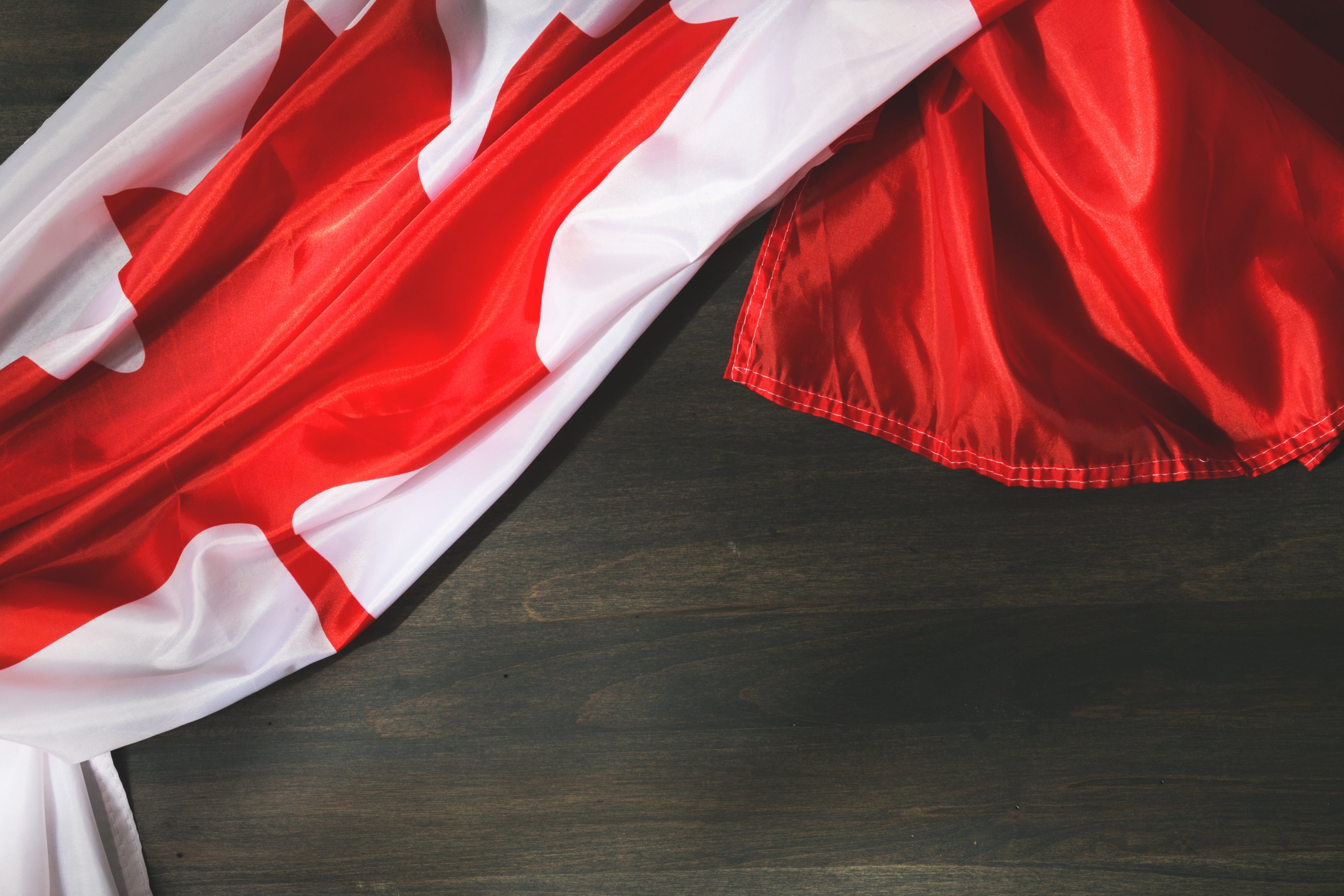 The bright red and white of the Canadian flag is gently piled on a smooth woodgrain surface. The light shines on the folds of the smooth flag texture.