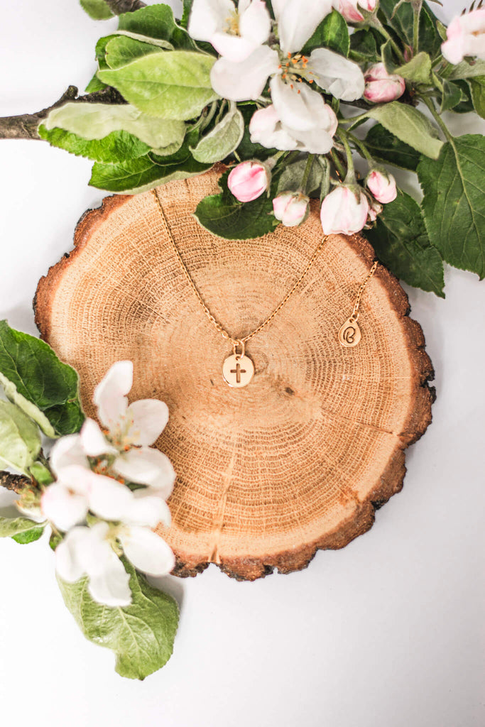 Srebrny naszyjnik celebrytka z okrągłą zawieszką z wyciętym krzyżem. To zawieszka o religijnej symbolice. Choker leży na ściętym pniu drzewa wśród kwiatów jabłoni.