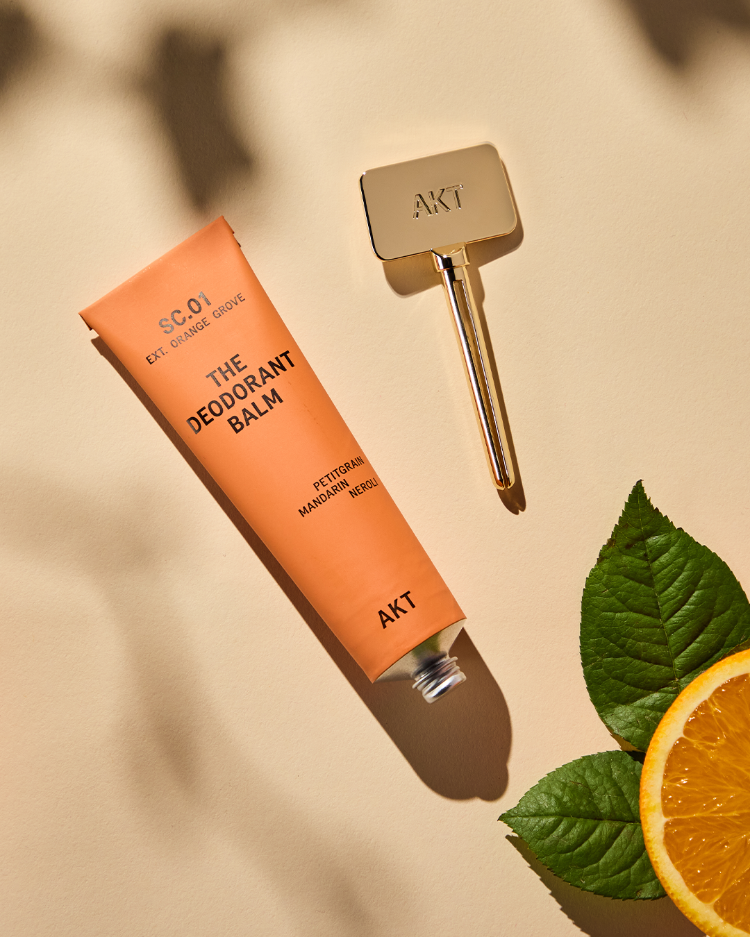 Deodorant balm tube with applicator, mint leaves, and a slice of orange on a beige surface.