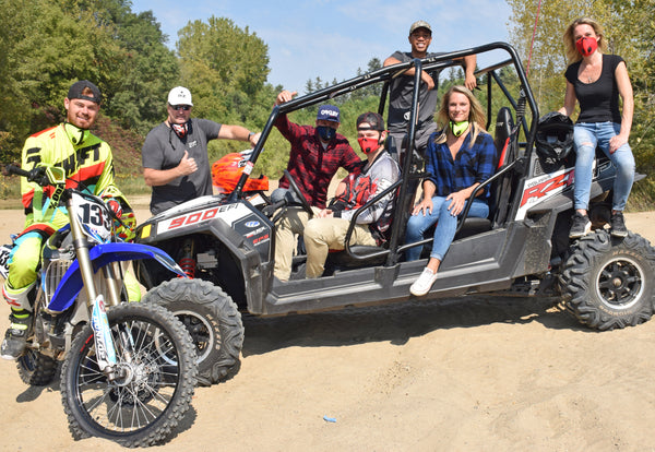 Group of men and women standing around a UTV and two dirtbikes wearing RZ masks for protection against dust and dirt on a sunny summer day. The individuals appear relaxed and happy, wearing casual outdoor clothing.