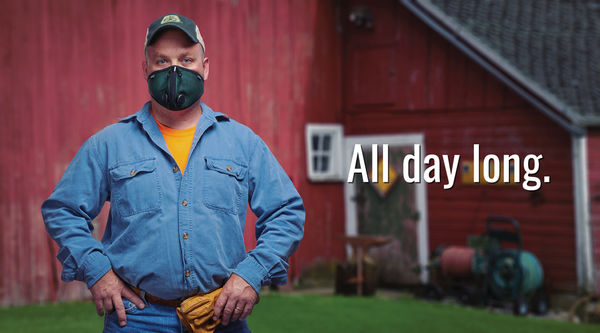 A rugged male farmer stands proudly in front of a barn, wearing an RZ Mask. He wears a plaid shirt and denim jeans, with work boots completing his outfit. The RZ Mask covers his nose and mouth, protecting him from dust and other particles while working in the farm. In the background, a classic red barn with a pitched roof can be seen, indicating a traditional farming setting. The farmer appears confident and determined, suggesting he is ready for any task at hand.