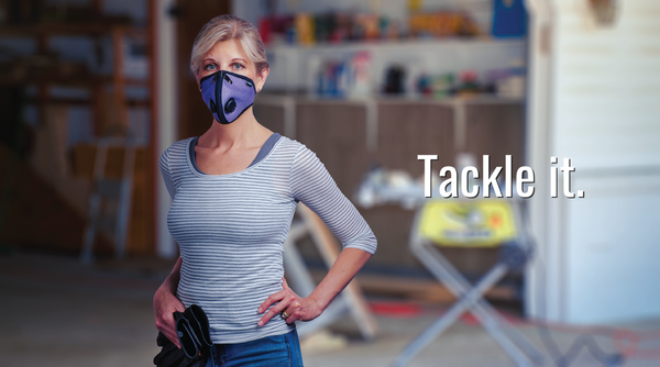 An active woman stands confidently in a garage, wearing an RZ Mask. She wears a casual outfit, including a light-colored t-shirt and dark pants. Her hair is tied back in a ponytail, and she appears ready for a project or task. The RZ Mask she wears covers her nose and mouth, providing protection from dust and other particles. In the background, various tools and equipment are visible, suggesting she is in a working environment.