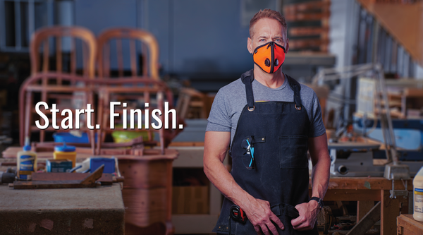 A skilled male woodworker stands in a woodshop, wearing an RZ Mask and an RZ Apron. He wears protective goggles and ear defenders, indicating that he takes safety seriously. His clothing consists of a plaid shirt and dark pants, with work boots completing his outfit. The RZ Mask and RZ Apron protect him from dust and debris while working with wood. In the background, various woodworking tools and equipment are visible, suggesting he is in the middle of a project. The woodworker appears focused and determined, highlighting his passion for the craft.