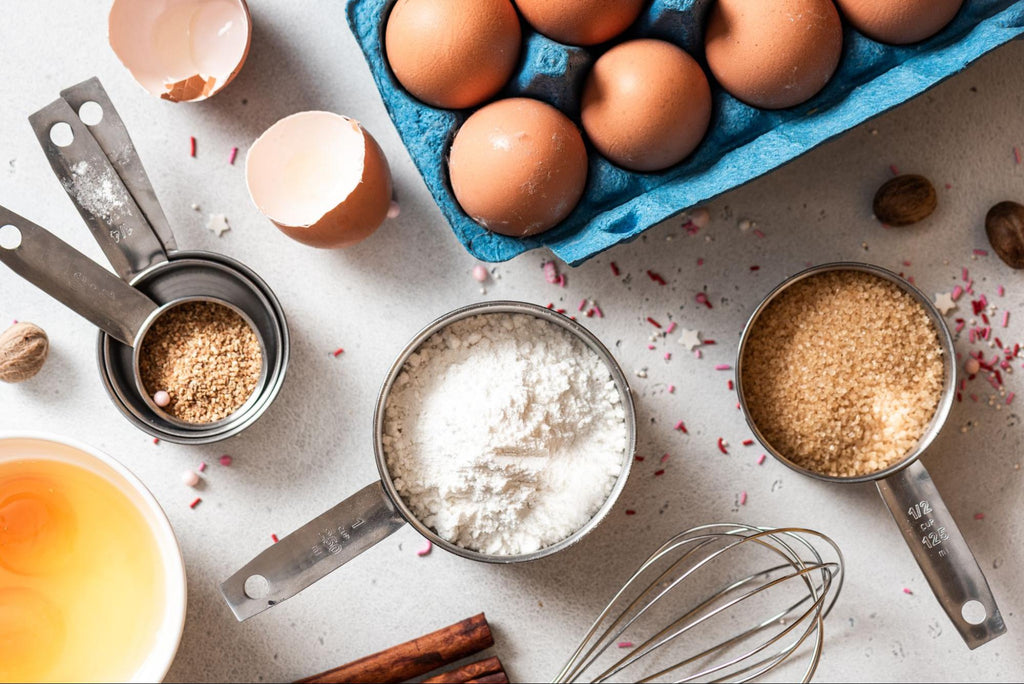A flatlay of pantry staples including brown eggs, flour, sugar, measuring cups, and a whisk. 