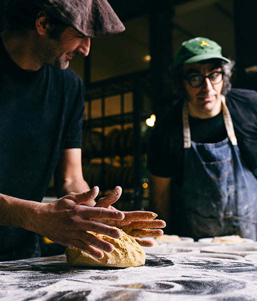 One person works dough on a flour covered table while another person looks on