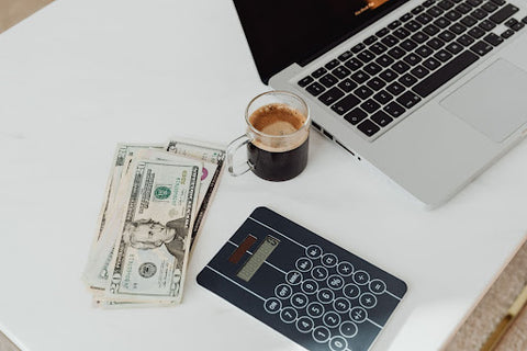 A stack of dollar bills, office calculator, coffee, and laptop laid on a white table