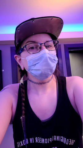 White woman from the waist up wearing a black vest with white writing on it. She is wearing a blue surgical mask and a black trucker hat with a titanium plate on the underside of the visor. She has brown hair coming down from behind her right ear in a braid, the braid has a silver cuff up by the ear and has a silver spike on the end of the braid A