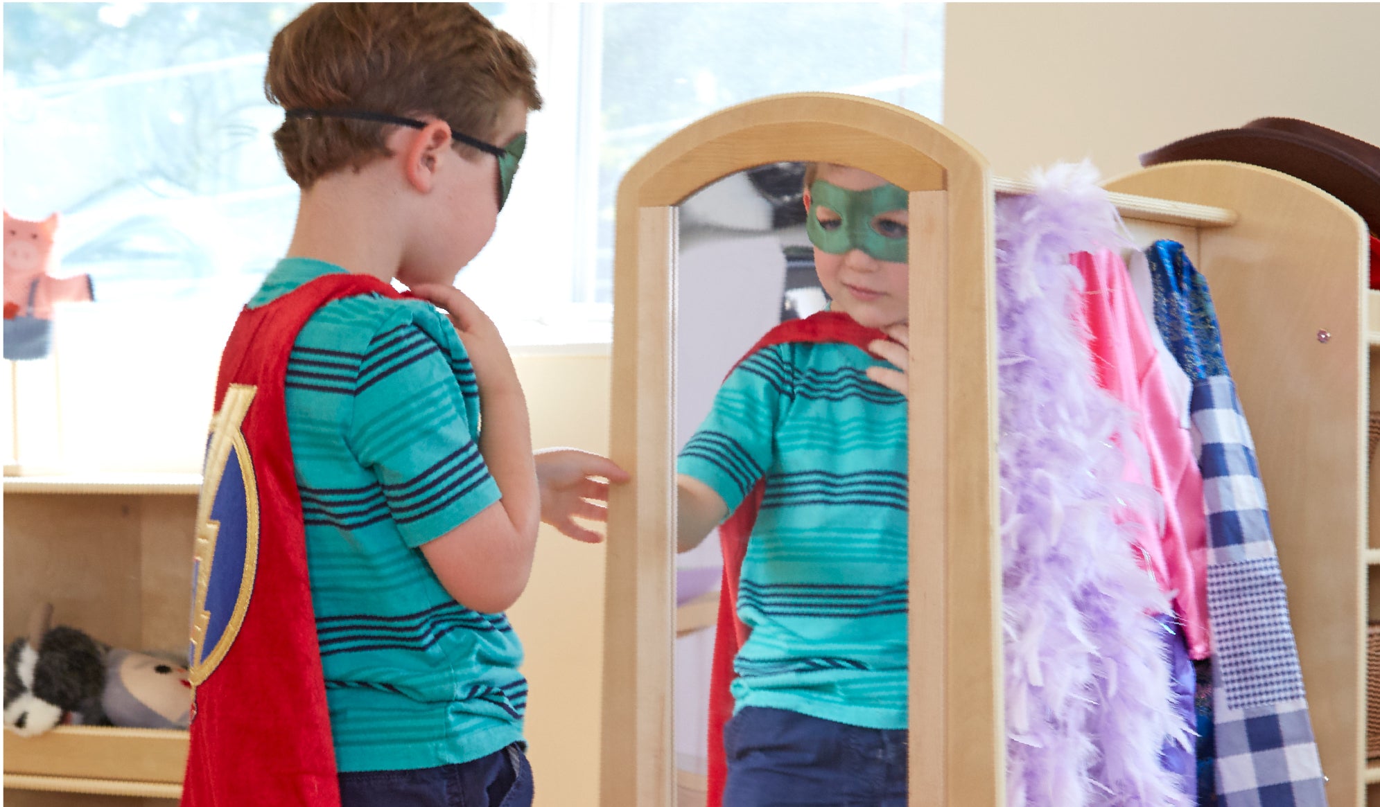 Image of a young boy looking at himself in the mirror of a Guidecraft dress up center dressed as a super hero.