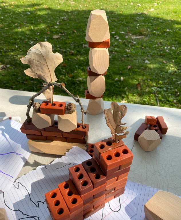 Image des petites briques Guidecraft et des pierres dressées Guidecraft Wood Stackers combinées à des pièces détachées naturelles comme des pierres, des bâtons et des feuilles dans un environnement de jeu extérieur.