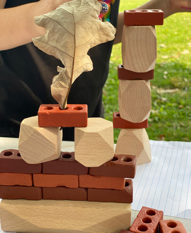 Imagen de pequeños ladrillos Guidecraft y apiladores de madera Guidecraft, piedras verticales combinadas con hojas en un entorno de juego al aire libre.
