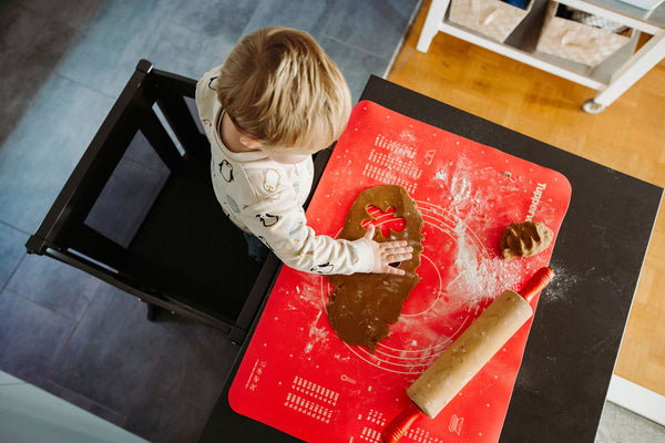 Young boy standing in Martha Stewart Kitchen Helper Toddler Step Stool - Charcoal, rolling out dough on red food mat at kitchen counter