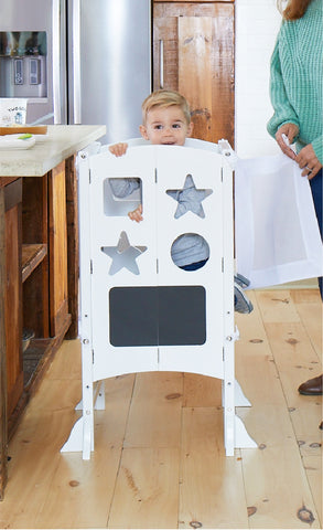 Image of toddler boy climbing into the Classic Kitchen Helper - White so that he can reach the counter and help his mother make breakfast.