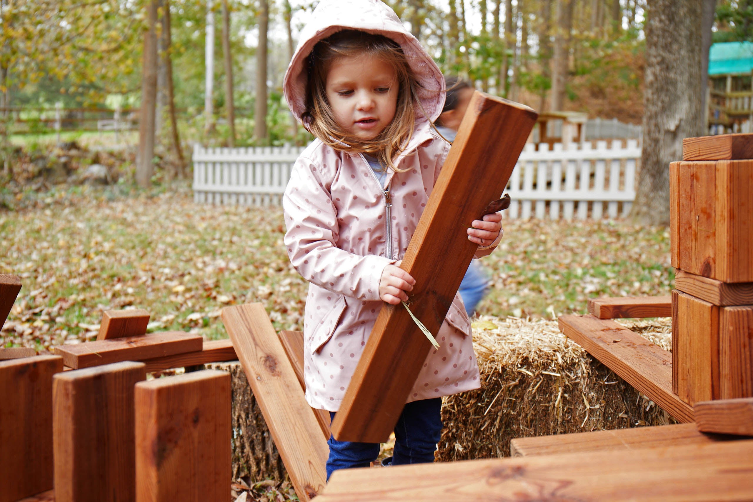 Image d'une jeune fille en imperméable jouant dehors avec des jouets de construction en bois.