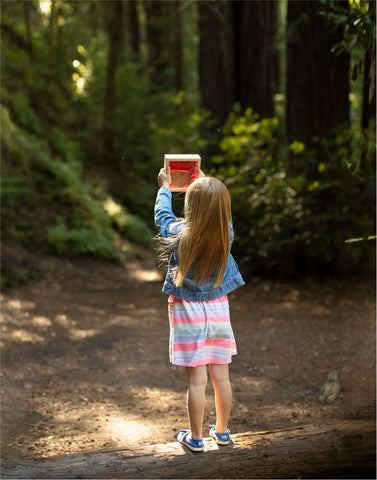 Image of a young blonde girl in the forest playing with the red piece from the Guidecraft Rainbow Stacking Pyramid set.