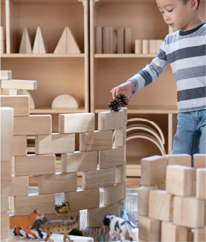 Image of a young boy in a classroom or playroom setting playing with Guidecraft Unit Blocks, natural loose parts and pretend play figures. Unit Blocks make the best holiday gift for kids 2 and up. 