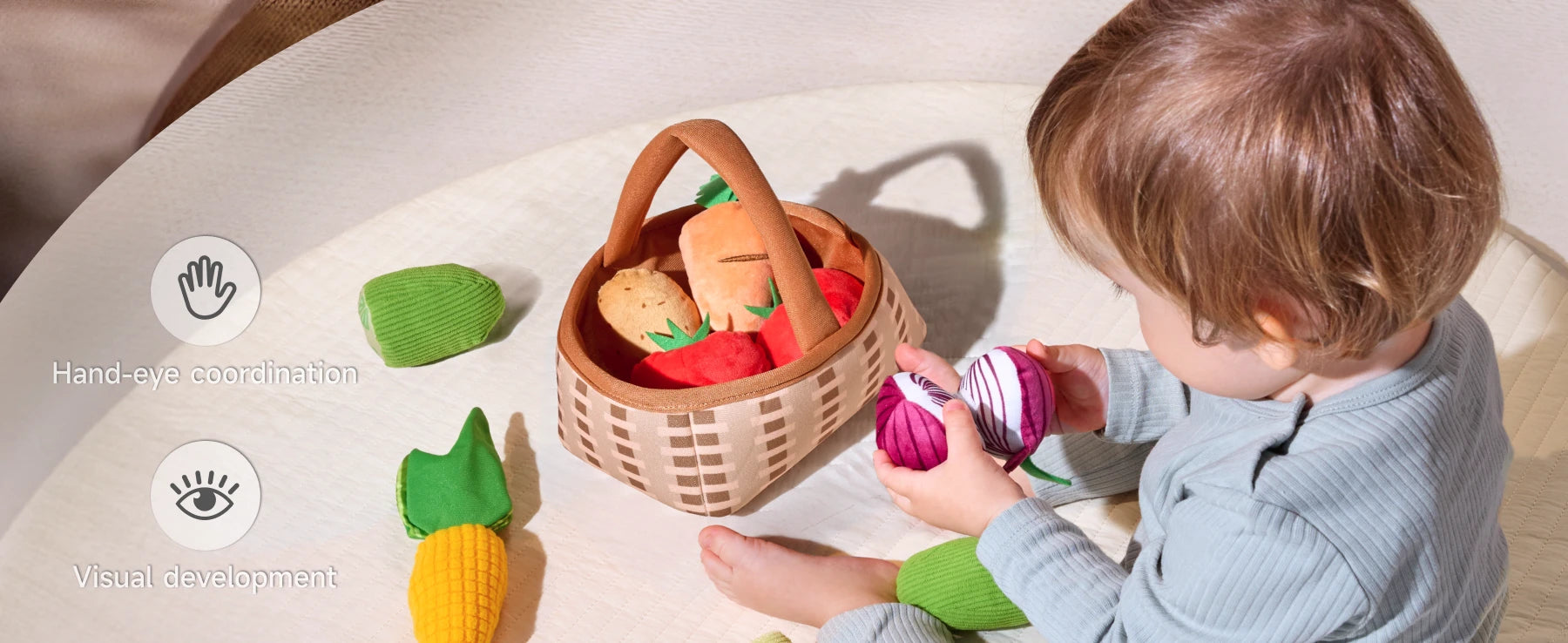 Baby with lively, felted vegetable toys—a strawberry, a carrot, a potato, and a corn cob—arranged on a textured surface beside a wicker basket.