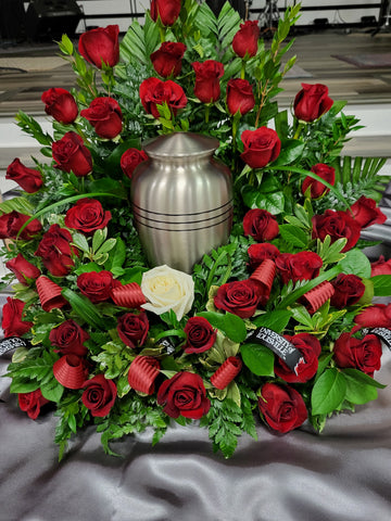 A silver urn sits inside of a floral wreath loaded with red roses. It has one white rose in the front with some red ribbon highlighting a local sports team.