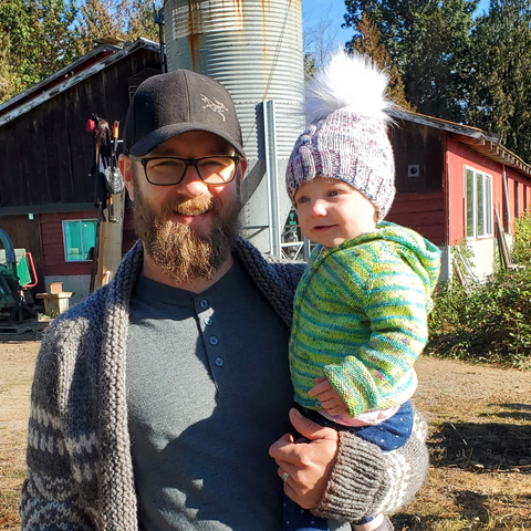 A bearded man holding a baby outside in the sun. Both of them are wearing hand-knit sweaters