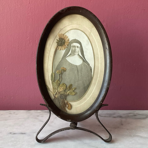 Metal oval frame containing a cabinet card of a nun and a pressed flower, circa 1900s-1910s.