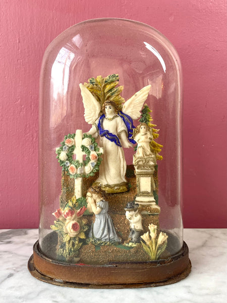Victorian diorama consisting of two-dimensional wax figures mounted inside a glass dome. An angel presides over two cemetery monuments and two weeping children.