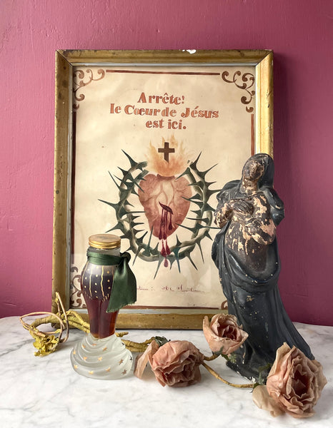 Vignette of antiques including a worn chalkware statue of the Madonna, a sacred heart watercolor painting, a Schiaparelli perfume bottle in the shape of a woman's lower half, and wax millinery flowers.