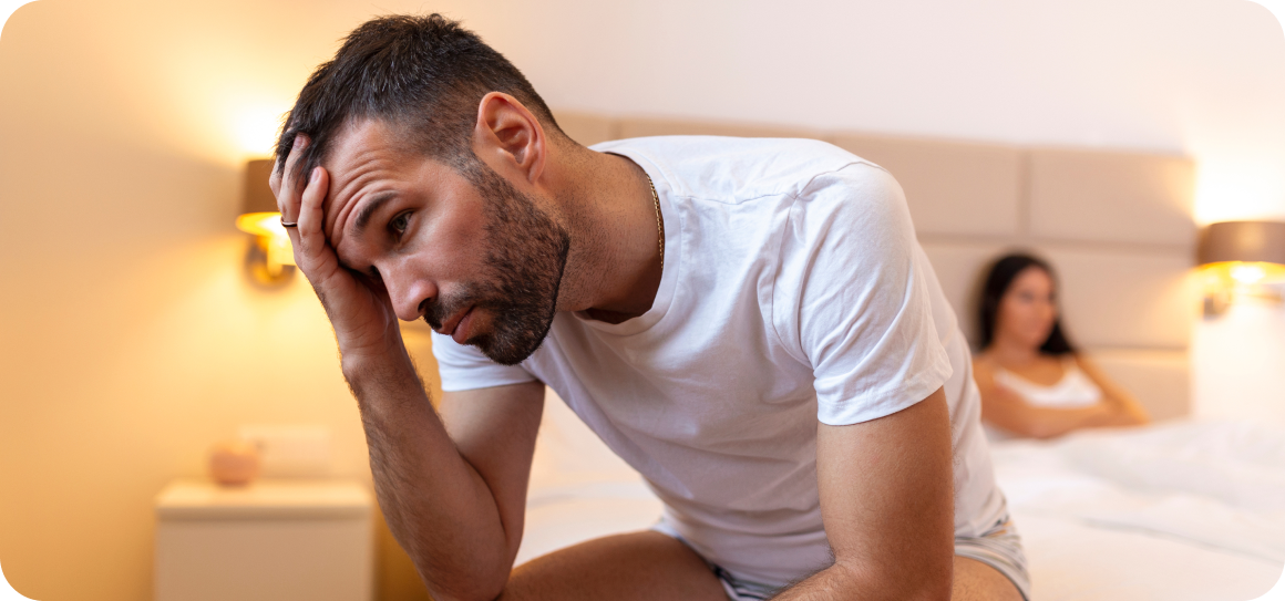 Man looking distressed, sitting on bed with woman in background.