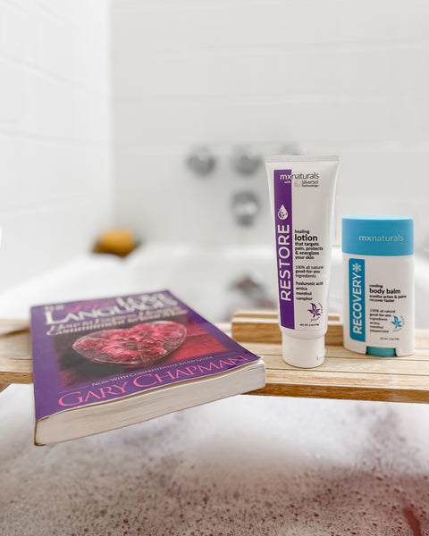 bubble bath with a tray across the tub holding a book called "The Five Love Languages" as well as mx naturals' restore targeted healing lotion and recovery cooling body balm.