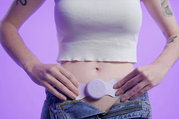 Person wearing white cropped tank top and unbuttoned blue jeans standing against purple background placing purple Aura TENS machine on lower stomach