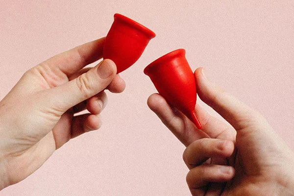 Two hands each holding one red menstrual cup against a peach background