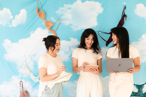 Three women of color with sensitive skin standing in front of a blue sky with clouds, dressed in workleisure outfits without bras..
