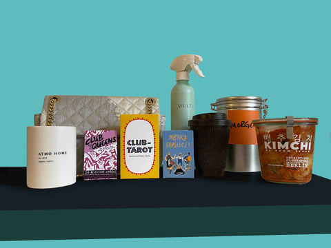 Photo of the following products from left to right: a white Atmo Home ceramic candle pot, a gray fabric purse with a golden chain, three card games: "Club of Queens", "Club Tarot", and "Merry Families," a green spray bottle that says "MULTI", a brown reusable coffee cup made from coffee grounds, an aluminum coffee can, and a glass jar of kimchi.