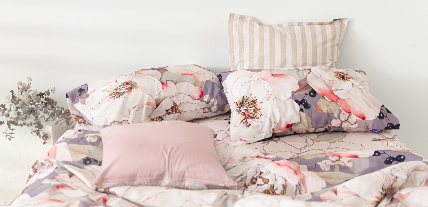 Parte superior de la cama, deshecha y con cojines, en tonos violetas y rosas con estampado floreado