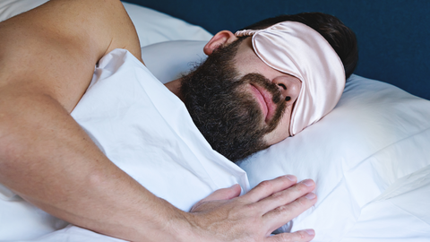 A man with a beard and sleep mask sleeping in a bed.