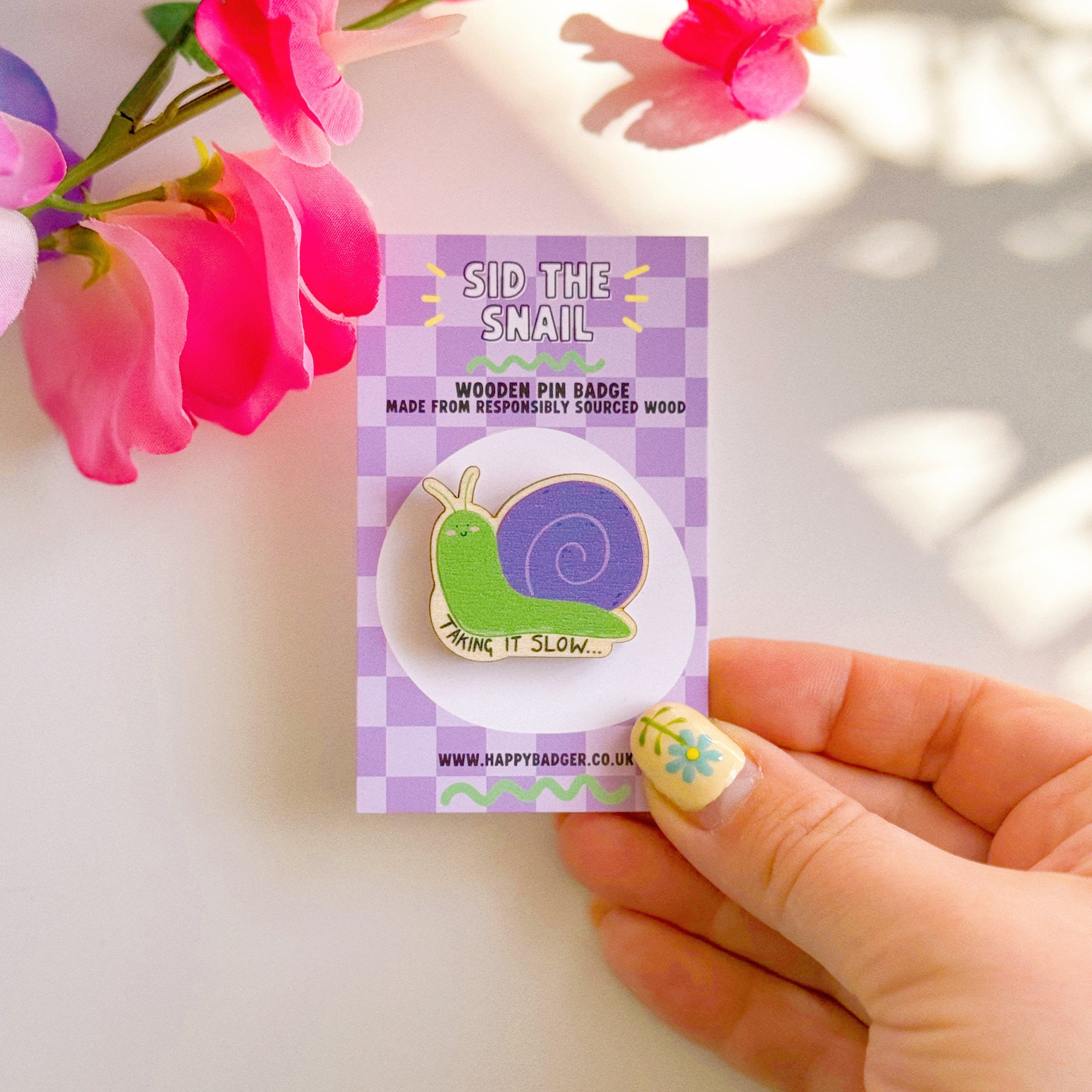 A cute wooden eco friendly pin badge, of a snail, that says 'taking it slow...'. The snail is purple and green and is smiling with rosy cheeks.