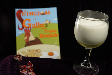 A glass of syllabub, which is a frothed cream desert, alongside a set of dice, with a book looming in the background.