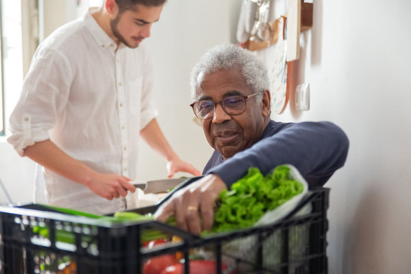 Senior man reaching for vegetables, while a male caregiver prepares a meal in the background.