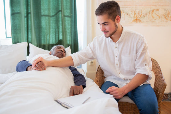 Man holding the hand of a senior man in bed while sitting next to it.