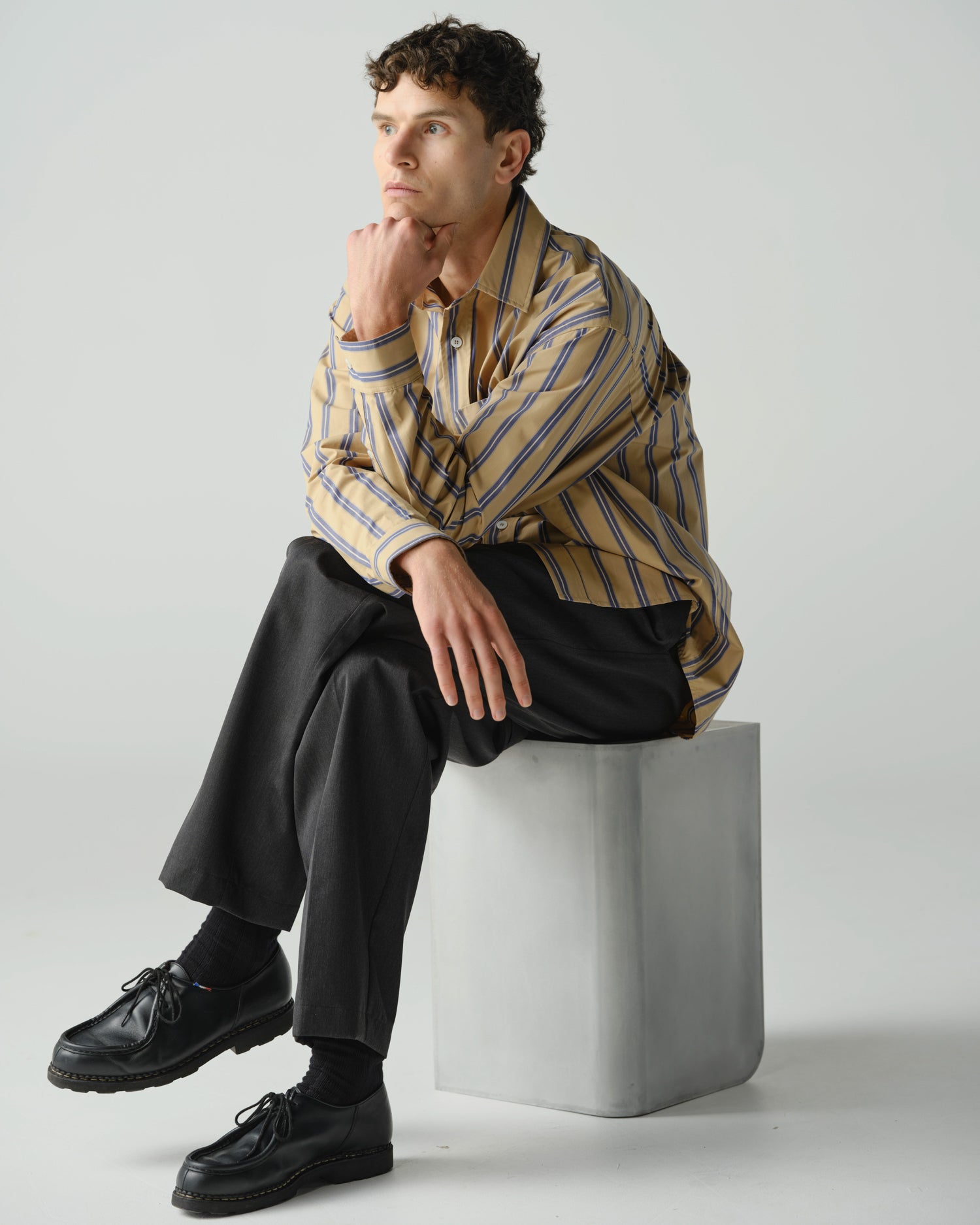 Man sitting on aluminium stool wearing a striped long sleeve shirt and charcoal tailored trousers.