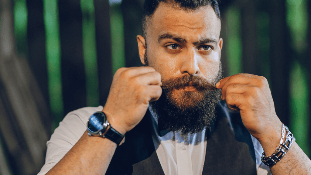 A confident man with a full beard and mustache, adjusting his mustache while wearing a suit and a wristwatch.