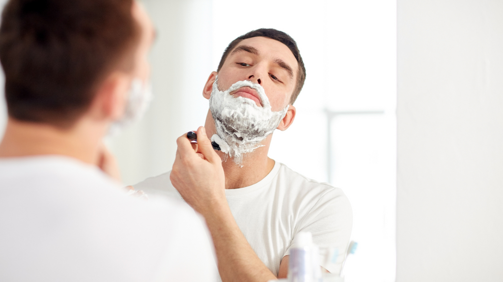 A man in a white t-shirt applying shaving cream to his face in preparation to shave his beard and mustache.