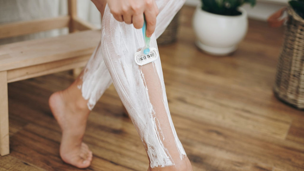 Person shaving their leg with foam using a handheld razor in a home environment, with wooden floor and a plant in the background.