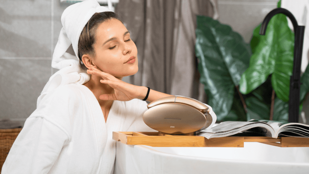 Relaxed woman in a bathrobe enjoying a facial treatment with JOVS Slimax device next to a bathtub