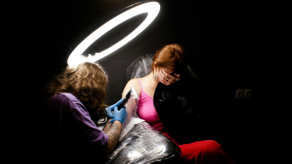 A person getting a tattoo on their arm by a tattoo artist under a circular light in a dark room.