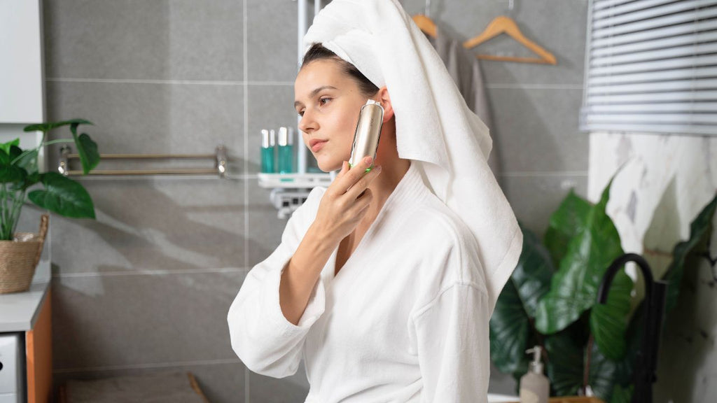 A woman in a white bathrobe is using a microcurrent facial device on her cheek.